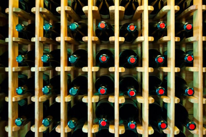 Botol-botol wine tersusun rapi dalam posisi miring di rak gudang penyimpanan yang sejuk dan remang.
