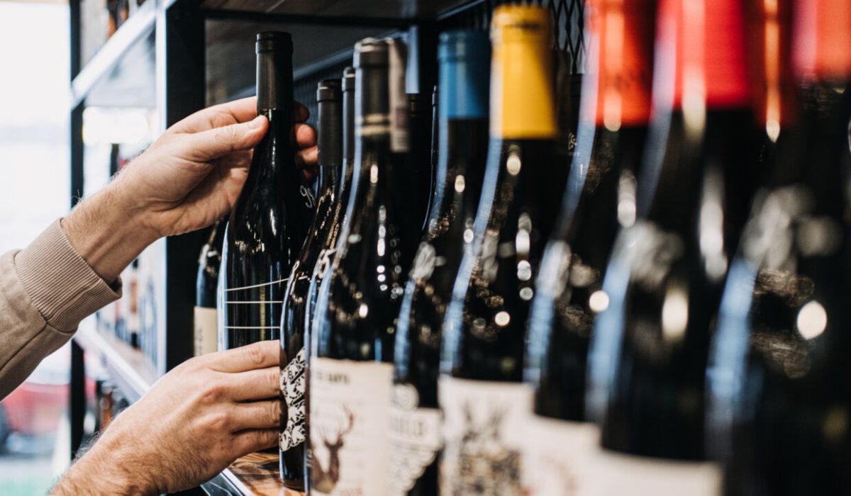Seorang pembeli sedang memilih sebotol wine dari rak yang dipenuhi botol-botol wine di sebuah toko.